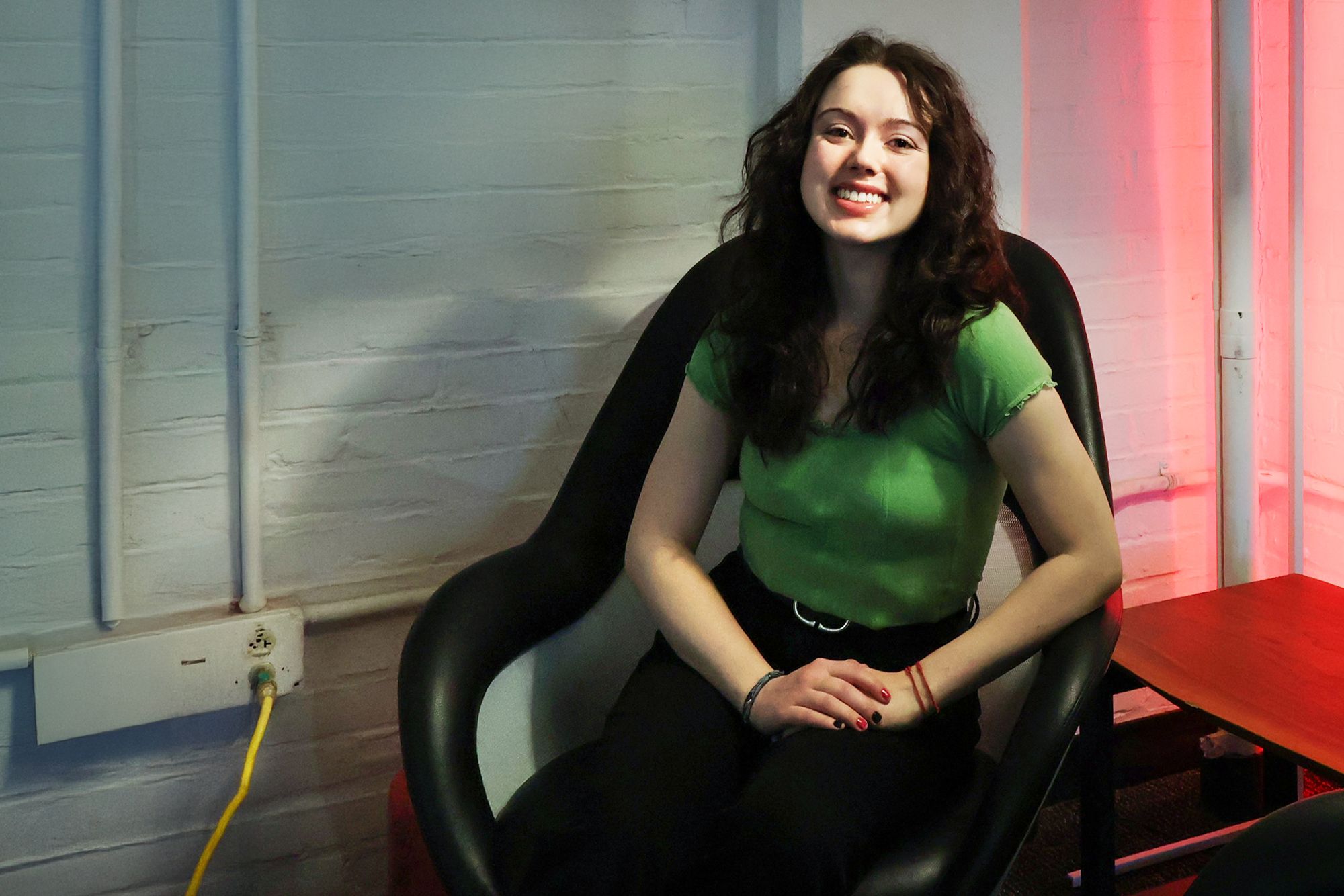 Young woman smiling and seated in a curved black chair against a white brick wall. She is wearing a bright green top and black pants, with red-painted nails and a red bracelet. The room is lit with a mix of white and red lighting, adding contrast to the scene.